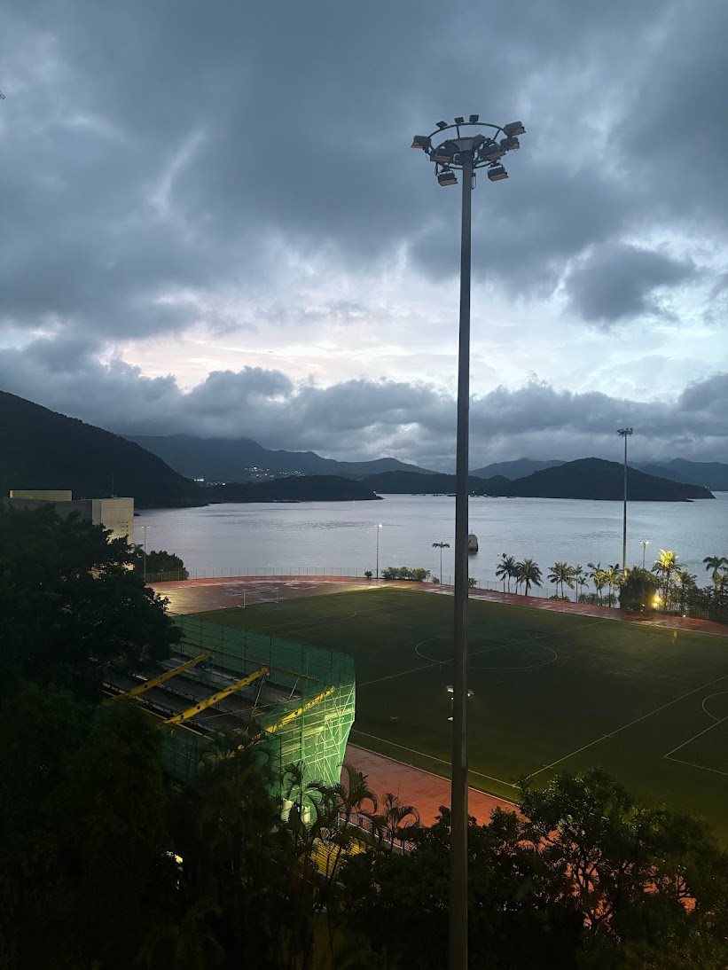 View over the HKUST sports field and Clear Water Bay at dusk, with mountains under a cloudy sky
