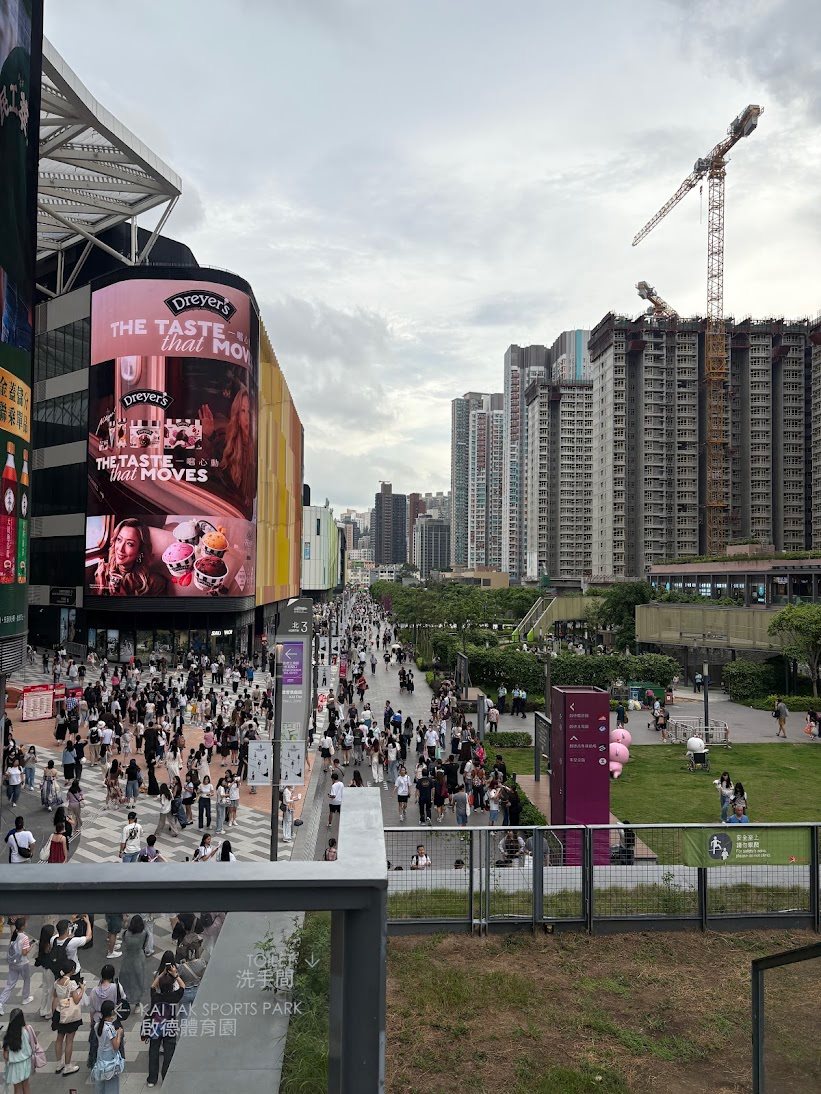 Large crowd walking along the promenade at Kai Tak Sports Park beside a curved video billboard and high-rise apartments